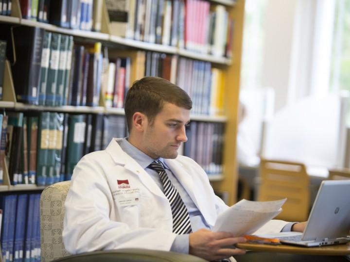 Student in white coat sitting in chair in front of the bookshelf doing work on their laptop